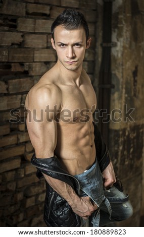 Muscular shirtless young man with jeans, indoors. Old brick wall behind. Looking at camera