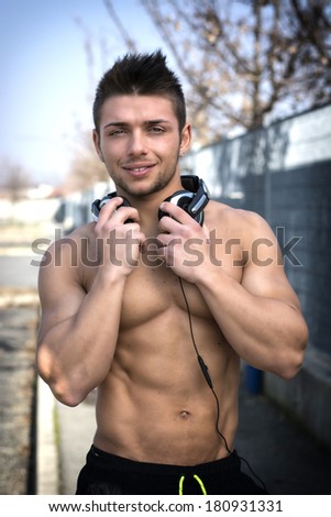 Muscular shirtless young man outdoors with headphones smiling and looking at camera