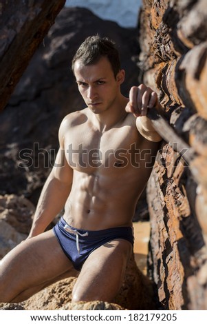Muscular shirtless young man sunbathing on rock by rusty metal wall, looking at camera