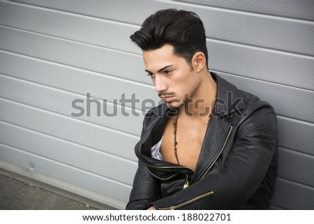 Young man with black leather jacket looking away, sitting on the ground outdoors
