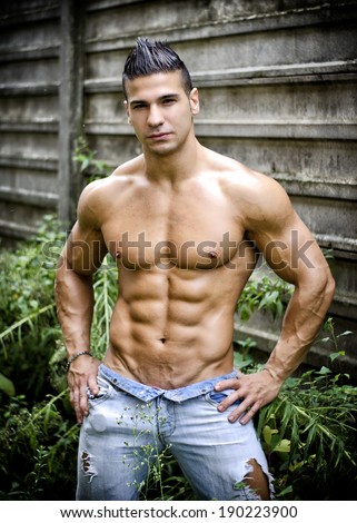 Muscular young latino man shirtless in jeans in front of concrete wall, with hands on hips