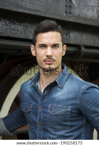 Handsome young man in denim shirt in front of old train, looking at camera