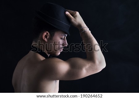 Profile of shirtless young latino man holding black fedora hat, on dark background