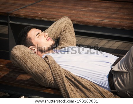 Attractive young man laying down on wood bench in the sun, eyes closed