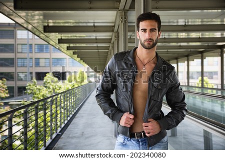 Handsome bearded young man standing, wearing leather jacket on naked torso, outdoors in urban environment