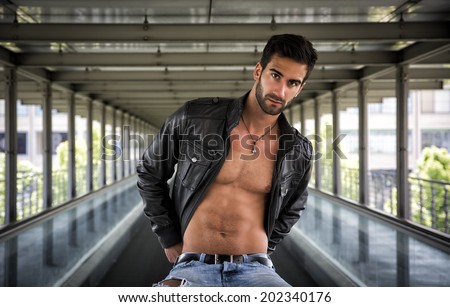 Handsome bearded young man sitting, wearing leather jacket on naked torso, outdoors in urban environment