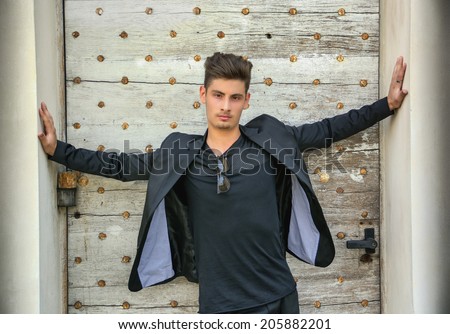 Handsome young man with arms spread open in front of old door, wearing elegant jacket, looking at camera