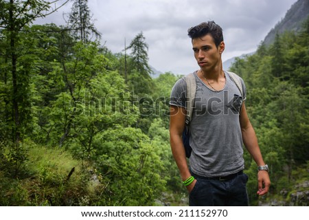 Handsome muscular young man backpacking or hiking in lush green mountain scenery pausing to look at something on the ground