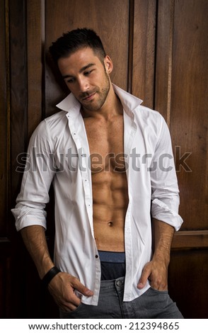 Sexy handsome young man standing in white open shirt with a smile in front of wood closet doors