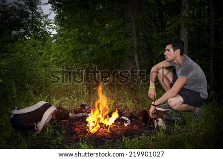 Young man camping in the mountains crouching alongside a burning campfire with his rucksack staring thoughtfully into the distance, profile view