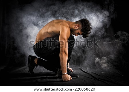 Handsome shirtless muscular young man kneeling down on black background, smoke around him. Looking down