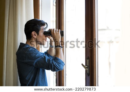 Young man standing looking through a glass door with binoculars as he watches something in the distance
