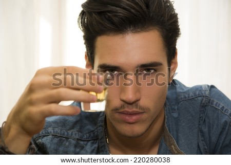 Seductive handsome young man sitting drinking toasting and celebrating with liquor in a shot glass as he gazes at the camera