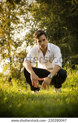 Handsome sexy man outdoors in the garden crouching down on fresh green grass amongst trees looking seductively at the camera