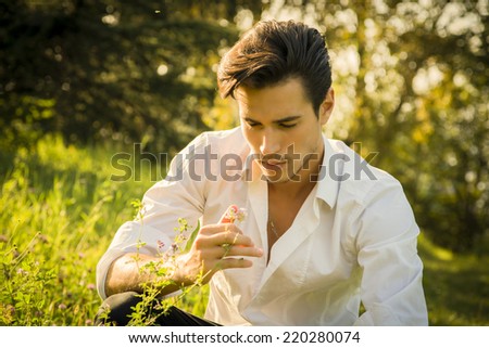 Nostalgic young man dreaming or reminiscing in the park as he kneels in green grass touching a flower with a look of wistful loneliness