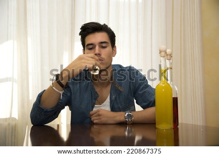 Young man sitting drinking alone at a table with two bottles of liquor alongside him sipping from shot glass to drown his sorrows