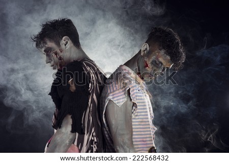 Two male zombies back to back on black smoky background, looking down and away