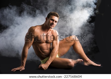 Attractive shirtless muscular man sitting on the floor in bathing suit on dark background