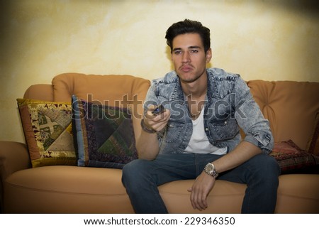 Unhappy, bored, unsure young man with remote control sitting on sofa, watching TV at home