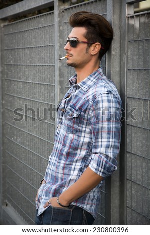 Trendy cool young man standing outside smoking, leaning against metal gate, wearing shirt and jeans