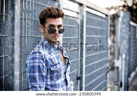 Trendy cool young man standing outside smoking, leaning against metal gate, wearing shirt and jeans