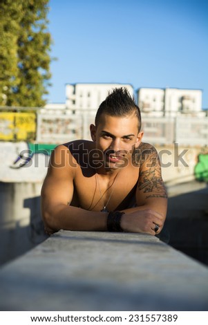 Handsome fit athletic shirtless young man in city setting leaning on concrete, looking at camera smiling