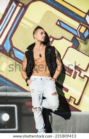 Handsome young man with shirt open on naked torso, standing against graffiti covered wall
