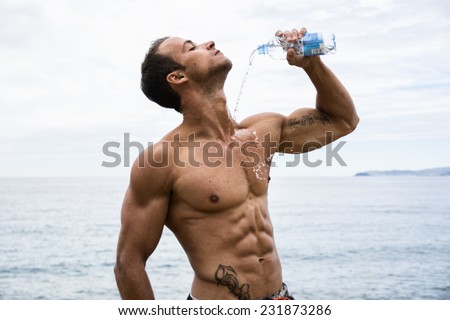 Attractive shirtless muscleman on the beach pouring down water on his chest from plastic bottle