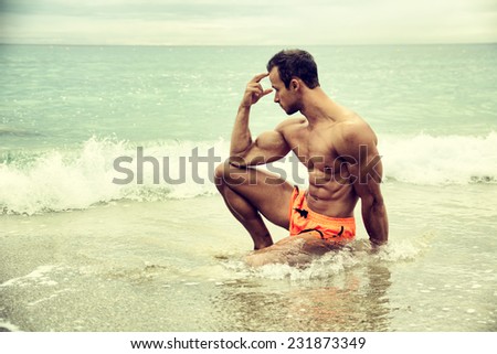 Handsome muscular young man on the beach sitting in the sea in profile pose