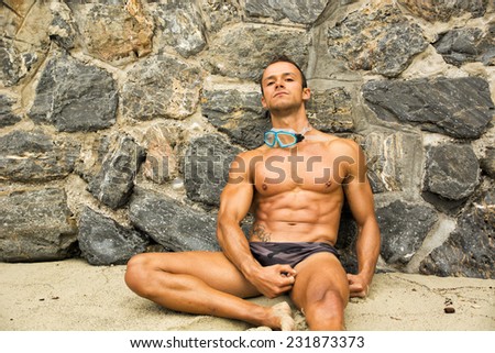 Solitary muscular young man leaning on a stone wall on the beach while listening to music through headphones