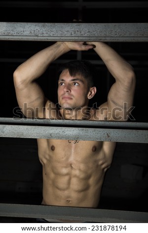 Muscular shirtless young man resting against metal structure, with arms raised above head