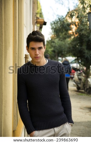 Handsome young man standing in European city street, leaning against a wall looking at camera