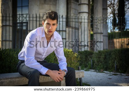 Handsome young man in European city, sitting on stone bench, looking at camera