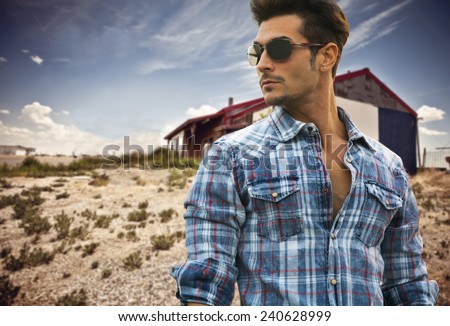Handsome fashionable man in sunglasses and a blue checked shirt posing outdoors with a wooden cabin behind and copyspace looking into the center of the frame