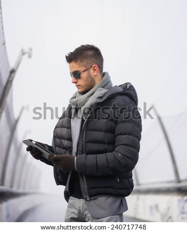 Handsome unshaven trendy man in winter fashion wearing a thick jacket, scarf and sunglasses standing looking down at a tablet computer that he is holding, side view