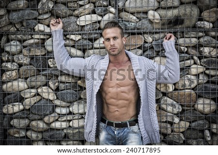 Muscular sexy man in front of stone wall, naked torso under knitted sweater, looking at camera