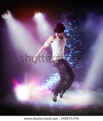 Trendy young man in hat hip hop dancing, breakdancing on a stage, in the beams of three spotlights leaping in the air with bent knees