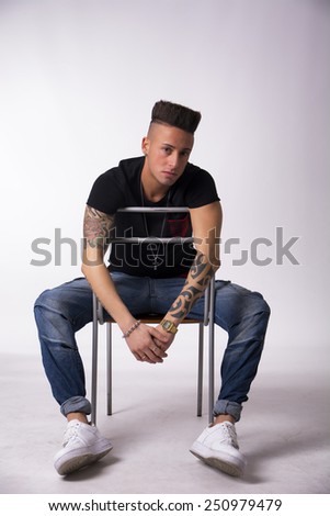 Trendy tattooed young man sitting on chair backwards, on light background