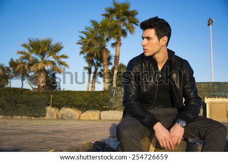 Fashionable handsome young man sitting waiting on a stone wall bordering a rural square or parking area with palm trees looking to the side watching