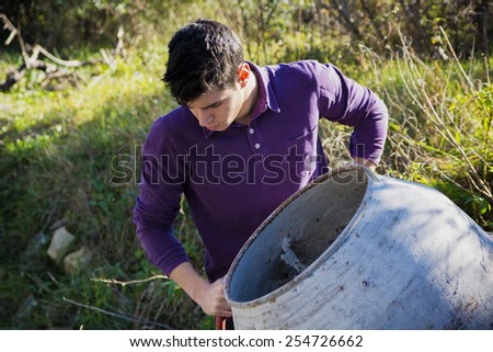 Handsome young man working with a cement mixer looking aside as he tilts the drum in a renovation or construction concept, high angle against long green grass