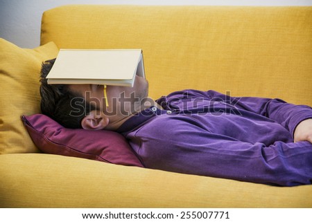 Over-worked, tired young man at home sleeping instead of working or studying, resting with head covered by open book