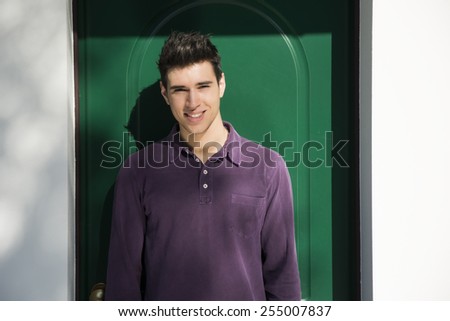 Handsome young man standing in front of house entrance, a green painted wooden door. Smiling at camera