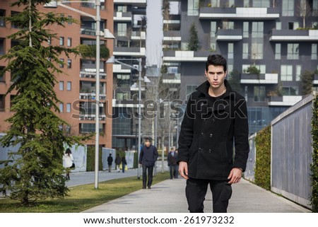 Stylish Young Handsome Man in Black Coat Standing in City Center Street with Skyscraper Behind Him, Looking to the Camera