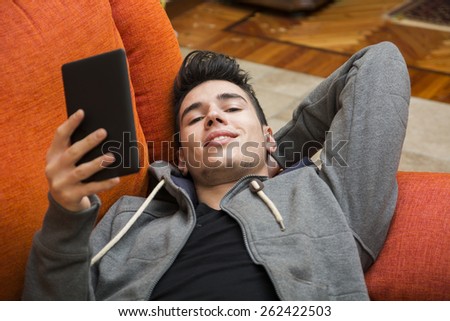 Handsome young man at home smiling, reading with ebook reader lying on a couch