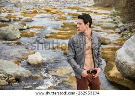 Handsome fit shirtless young man next to water pond or river, wearing grey leather jacket open on naked torso