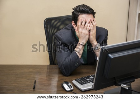 Desperate, worried young businessman sitting at his desk in front of his computer with his face in his hands and eyes closed