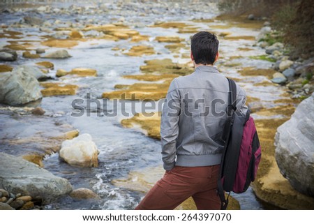 Attractive young man outdoor in nature, at river or water stream, seen from the back, with backpack or rucksack