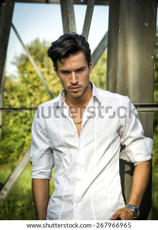 Handsome young man leaning against metal electricity trellis wearing white shirt, looking down