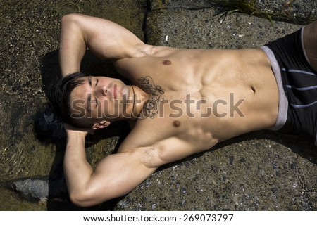 Handsome shirtless muscular young man sunbathing on rock near water in summer