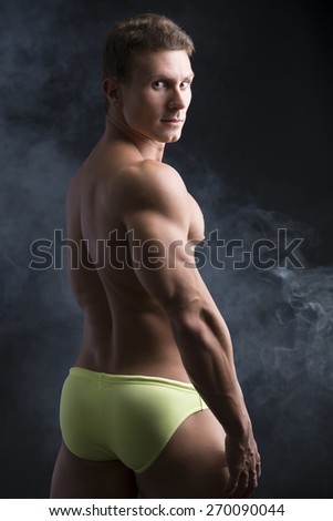 Handsome shirtless muscular man\'s back, turning around to look at camera, on dark background